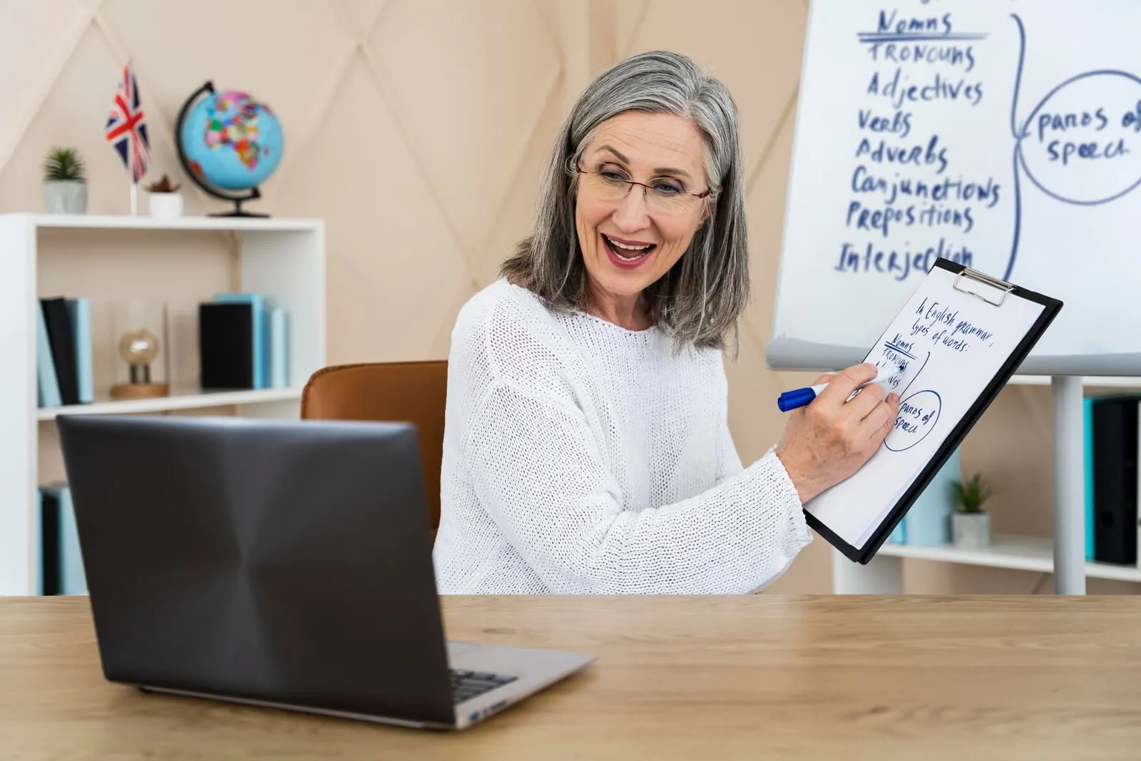 An older woman in glasses writing on a clipboard at a desk with a laptop, grammar notes on a whiteboard behind her