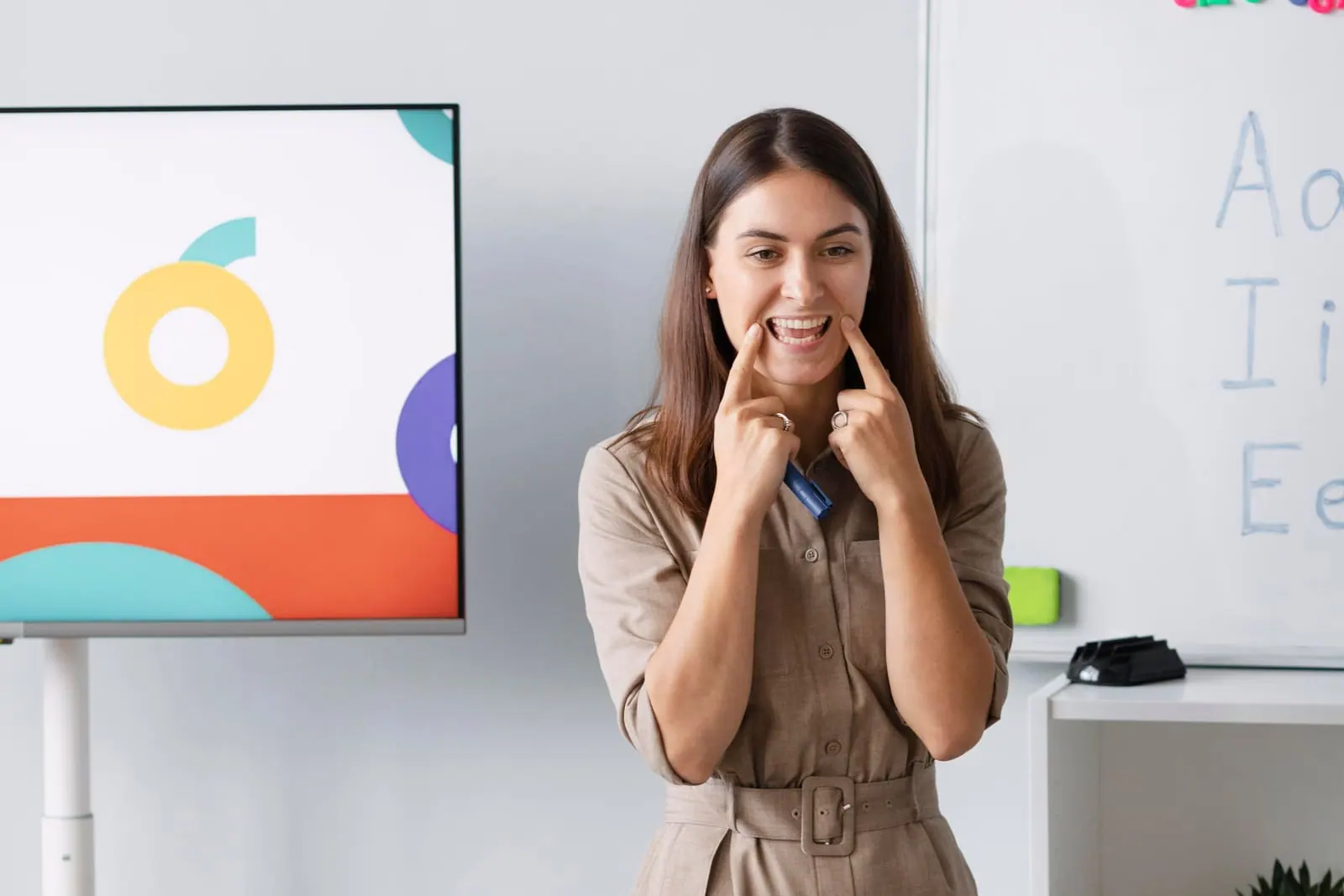 A teacher smiling in front of a classroom display and whiteboard while young students watch