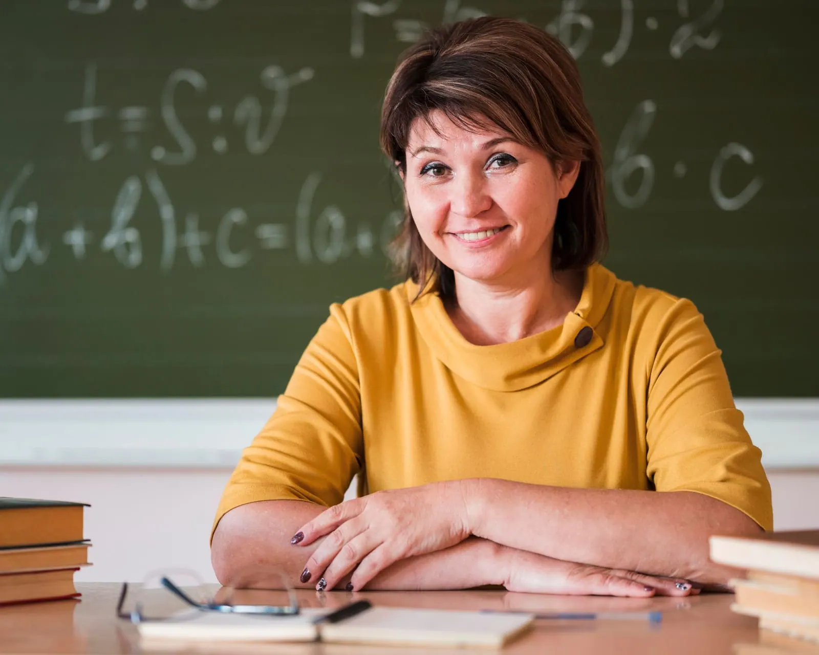 A smiling teacher seated in front of a chalkboard covered in equations