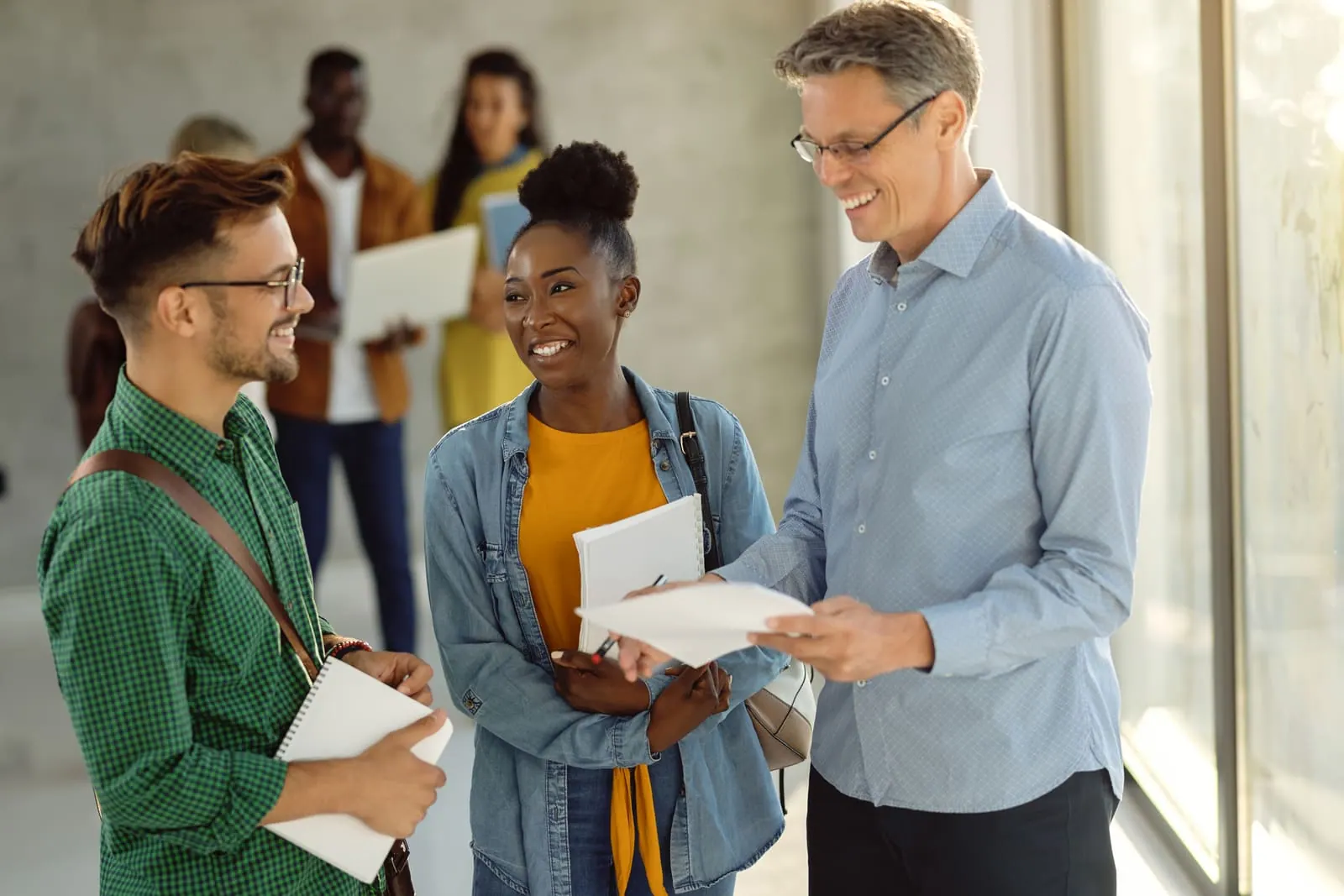 Three colleagues talking and smiling over a document in a bright campus hallway