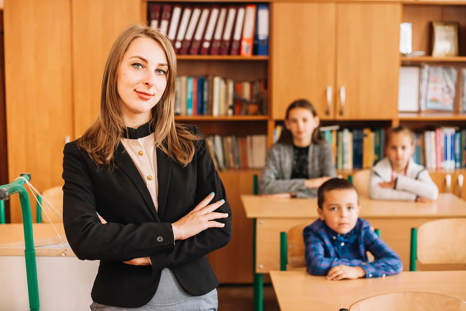 A woman in a blazer standing with her arms crossed in a classroom, with students seated at desks behind her