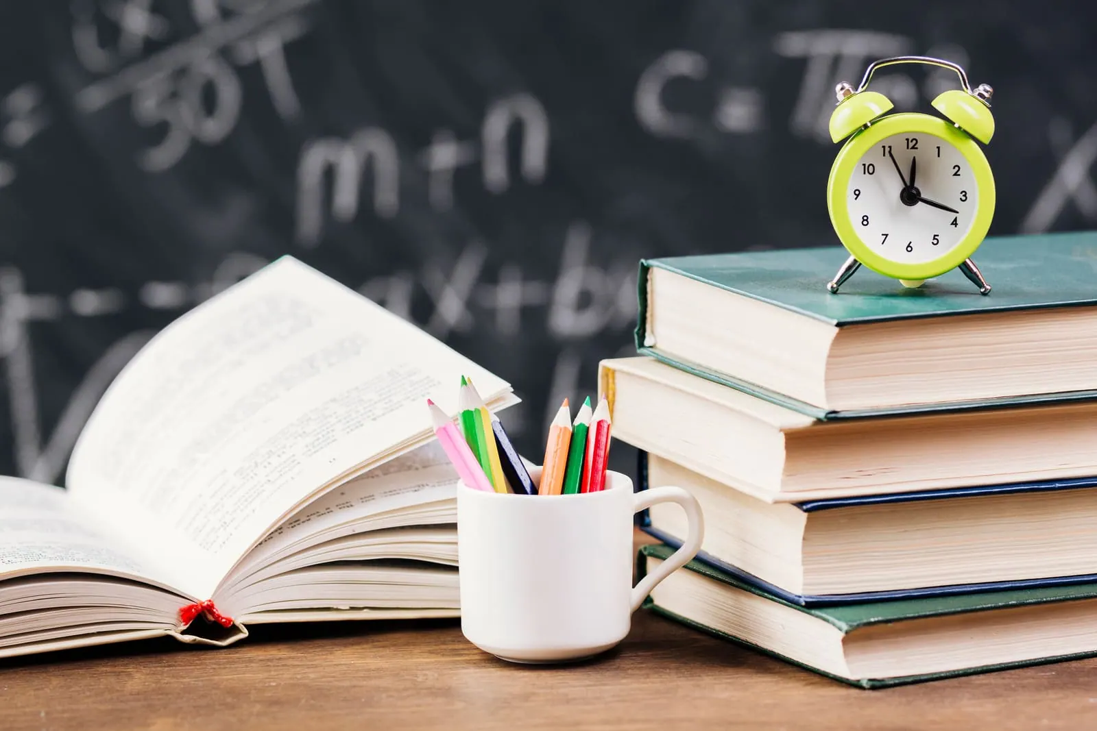 A stack of books, an open notebook and an alarm clock in front of a chalkboard