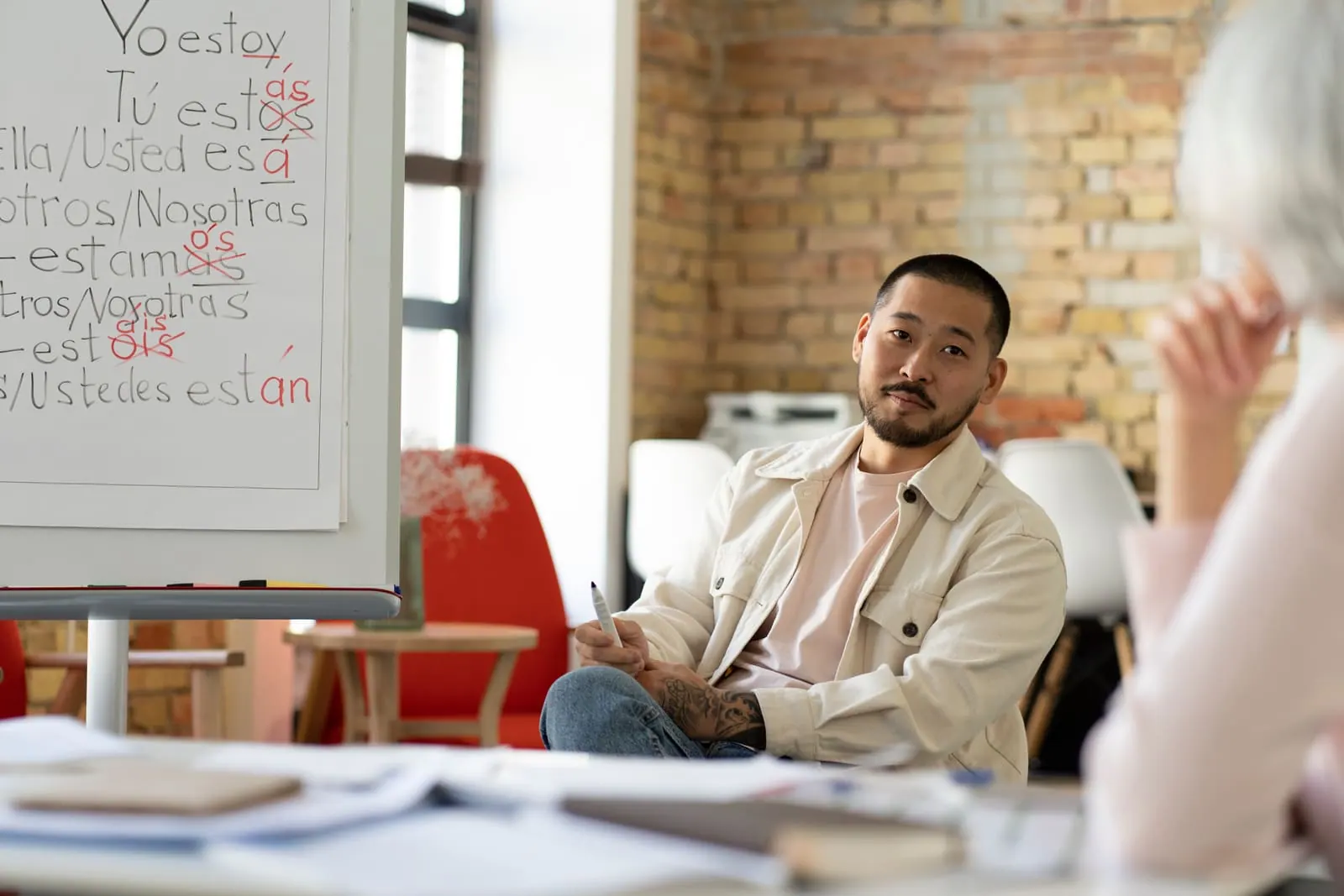 A man seated in a classroom beside a whiteboard of Spanish verb conjugations