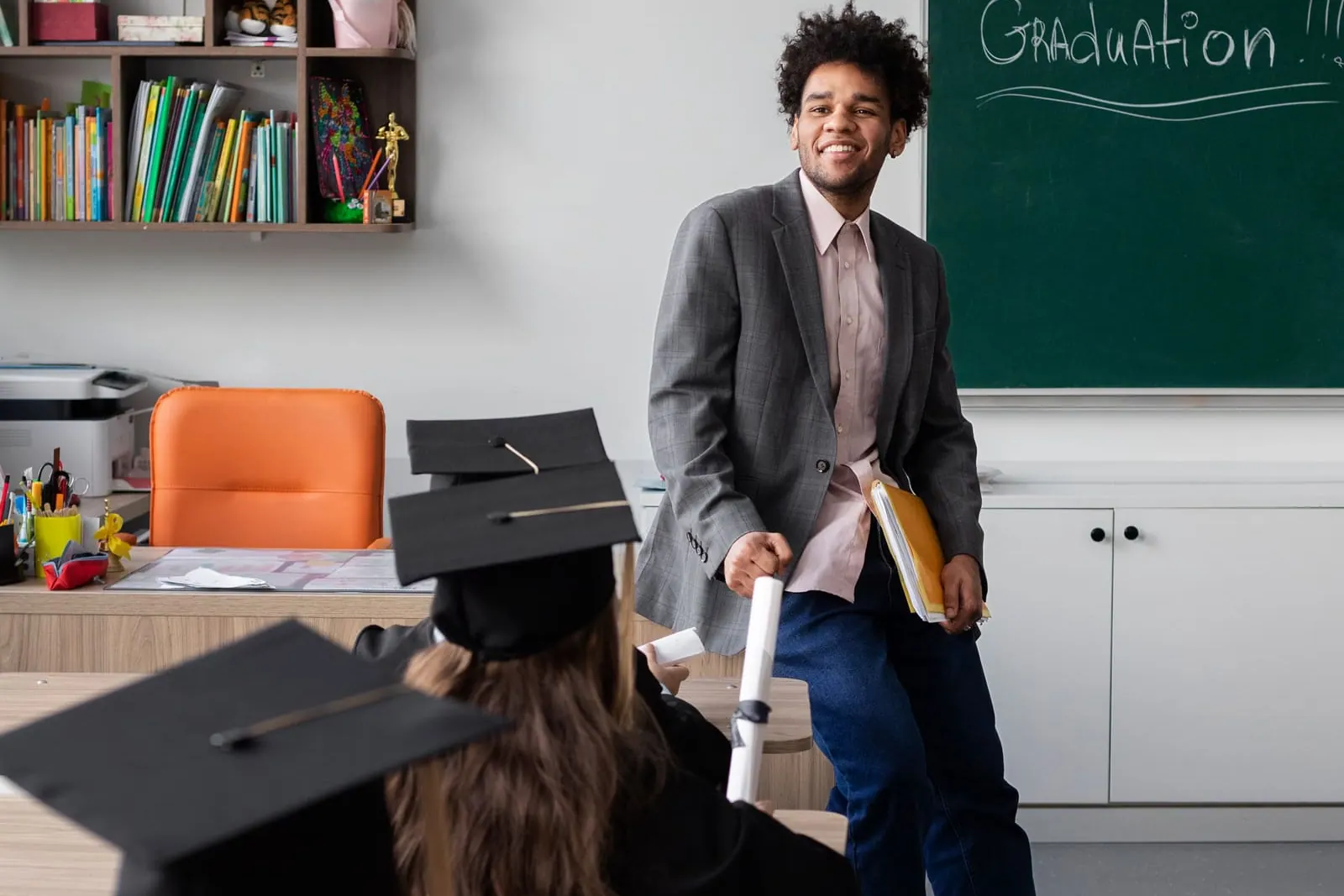 A young teacher standing beside a chalkboard marked Graduation while students in graduation caps sit at desks