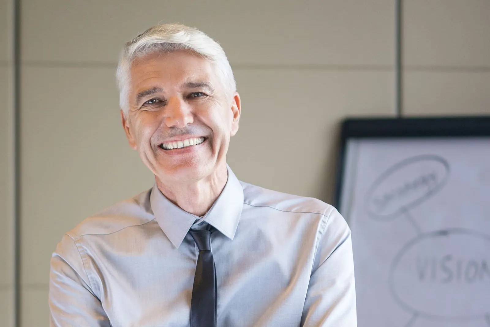 A smiling gray-haired man in a shirt and tie with his arms crossed beside a whiteboard